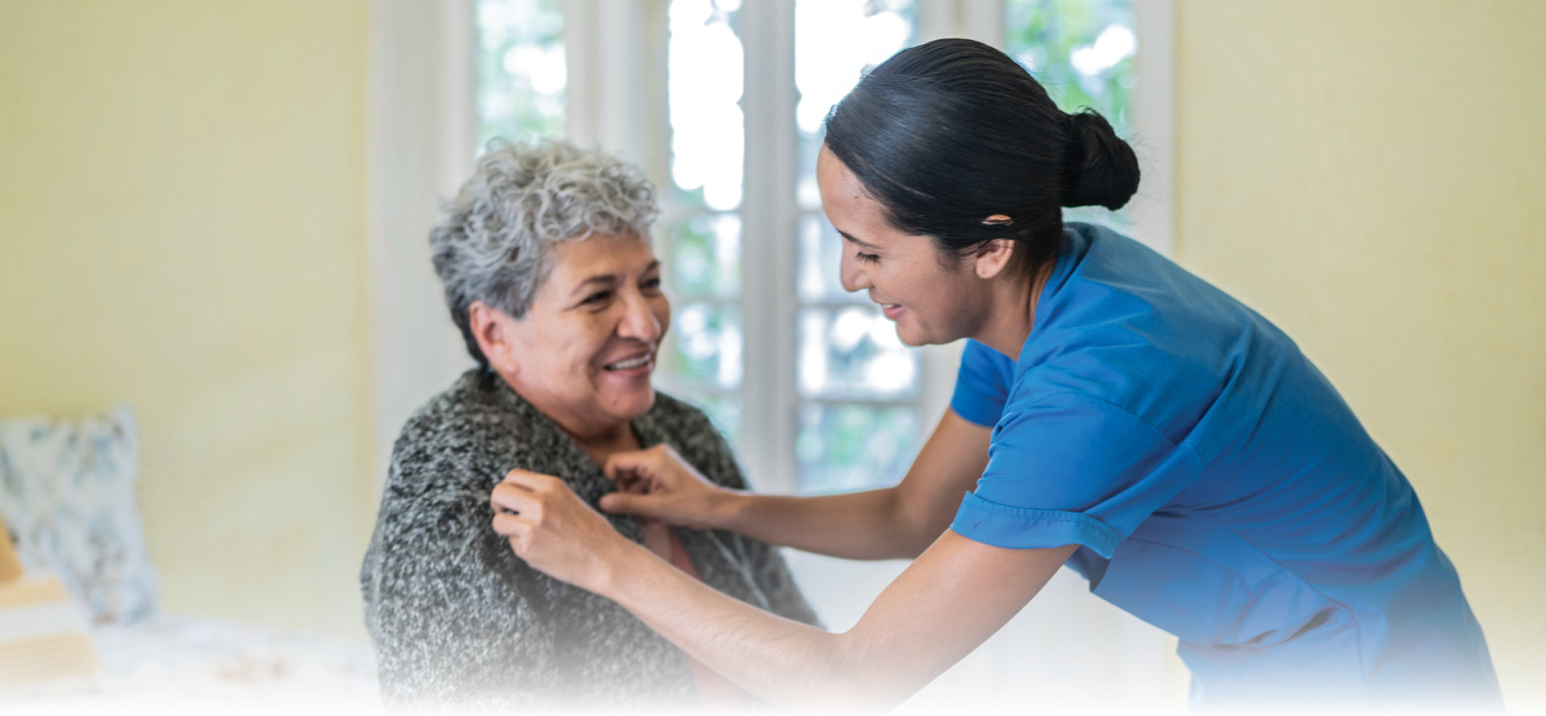 Adult nurse helping senior woman dress shirt in bedroom at nursing home